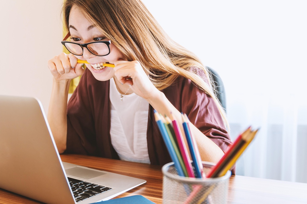 Lady at laptop Woman wearing glasses sat at laptop with pencil in her mouth and pencil pot on the desk